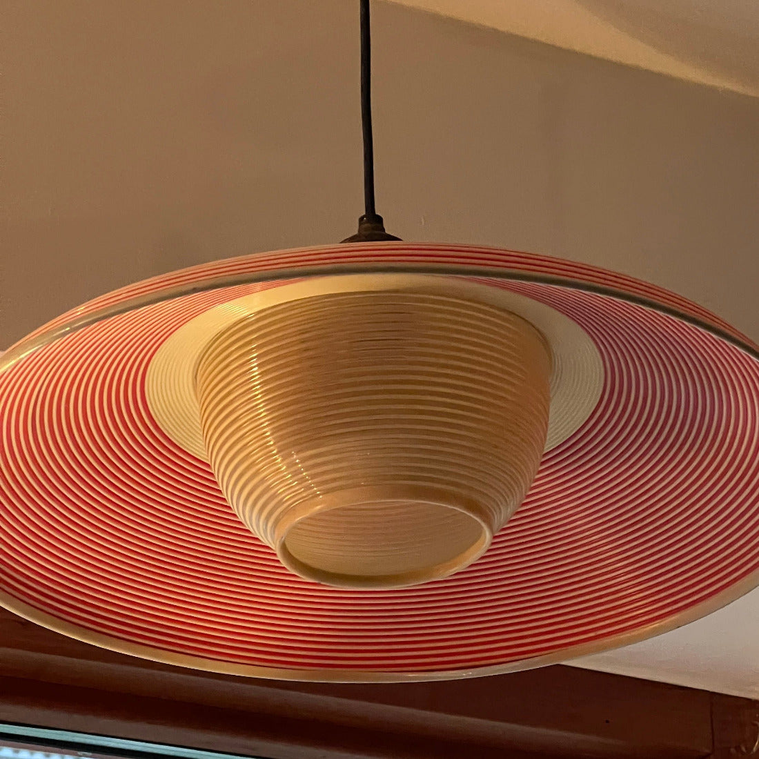 Lampskärmen är tillverkad i tålig plast med en karakteristisk randig design i rött och beige, kombinerat med mjukt cremevita detaljer som skapar en varm och inbjudande atmosfär. 