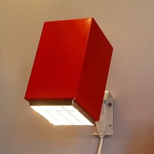 Vägglampan är tillverkad av rödlackerad plåt och har ett rutmönstrat raster i silverfärgad plast. 
